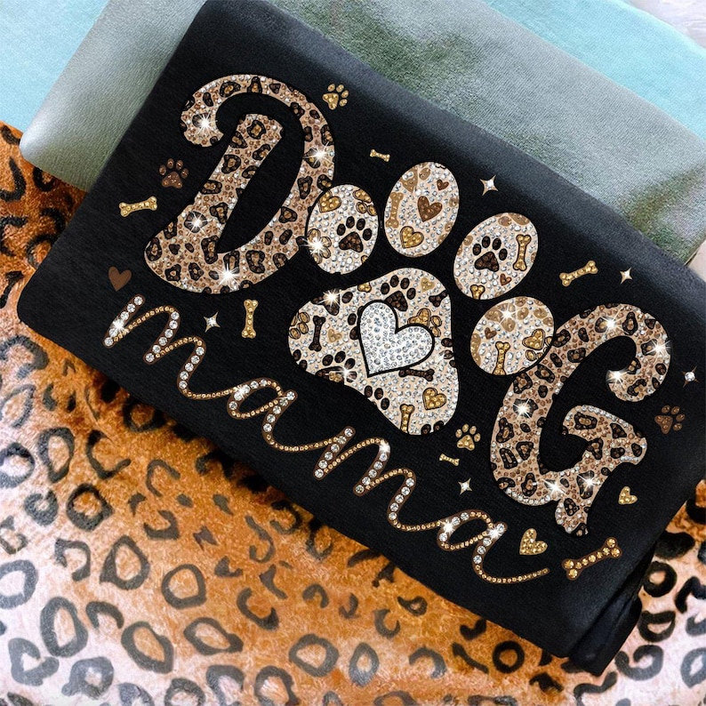 May include: Black t-shirt with the words "Dog Mama" in a sparkling, cursive font. The word "Dog" is in a leopard print design, and the paw prints have a heart and bone pattern. The shirt is folded on a leopard print fabric.