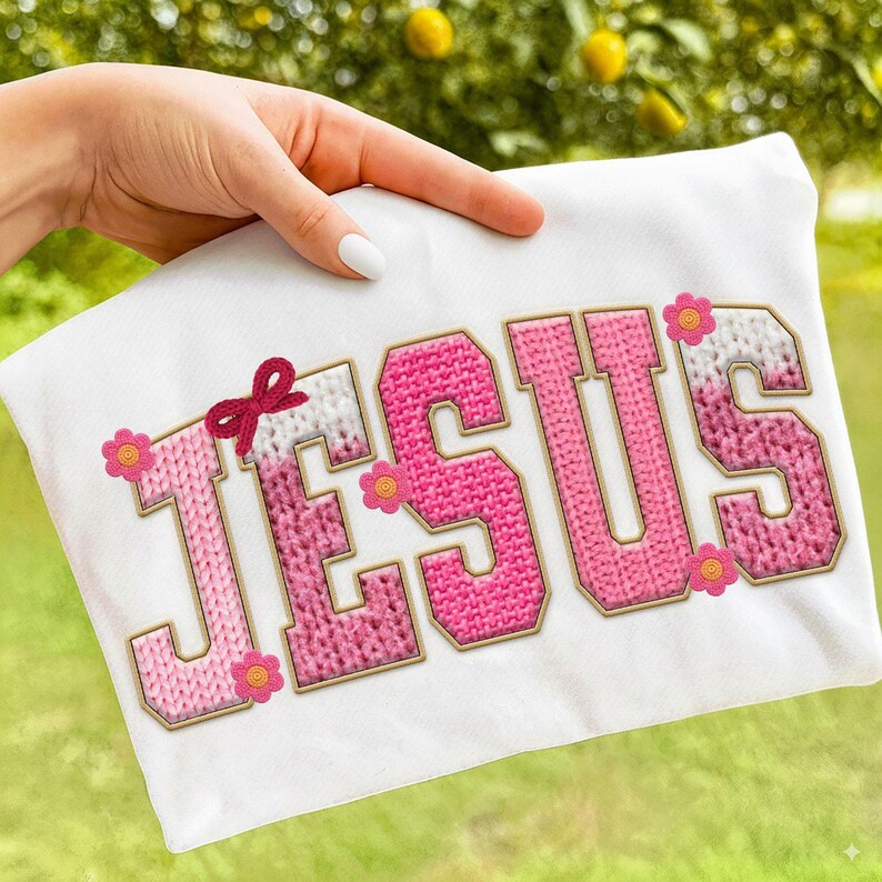 May include: White t-shirt featuring the word "JESUS" in pink and white knit-style letters with gold trim. A red bow and pink flowers accent the design. The shirt is held by a hand.