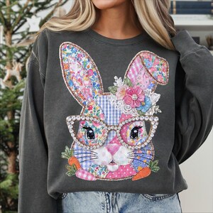 May include: A dark gray sweatshirt featuring a patchwork bunny design. The bunny has floral ears, a pink nose, and wears rhinestone-studded glasses. The design includes carrots and flowers, perfect for Easter.