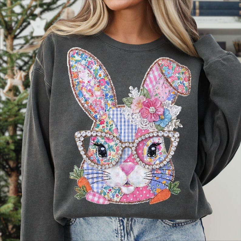 May include: A dark gray sweatshirt featuring a patchwork bunny design. The bunny has floral ears, a pink nose, and wears rhinestone-studded glasses. The design includes carrots and flowers, perfect for Easter.