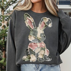 May include: Dark gray sweatshirt featuring a patchwork bunny design. The bunny is made of fabric in shades of pink, green, and beige, with a distressed, frayed edge. The sweatshirt has a crew neck and long sleeves, suitable for casual wear.