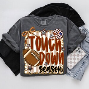 May include: Dark gray t-shirt with a football-themed graphic. The design includes a football, a helmet, and the words "Touch Down Season" in brown and white. Accents include stars and a bow.