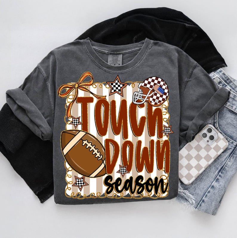 May include: Dark gray t-shirt with a football-themed graphic. The design includes a football, a helmet, and the words "Touch Down Season" in brown and white. Accents include stars and a bow.