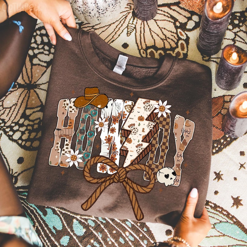 May include: A brown sweatshirt featuring the text "HONEY DAY" in a decorative style. The design incorporates a cowboy hat, flowers, a lightning bolt, and a rope bow. The sweatshirt is held by two hands.