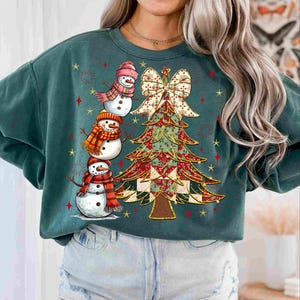 May include: A teal green sweatshirt featuring a festive Christmas tree and three snowmen. The snowmen wear hats and scarves, and the tree is adorned with a bow. Red and gold stars decorate the background, creating a holiday theme.