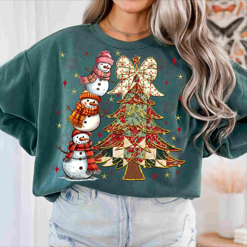 May include: A teal green sweatshirt featuring a festive Christmas tree and three snowmen. The snowmen wear hats and scarves, and the tree is adorned with a bow. Red and gold stars decorate the background, creating a holiday theme.