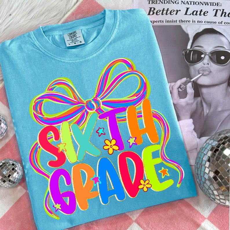 May include: A light blue t-shirt with the words "SIXTH GRADE" in colorful, bubble letters. The design includes a large, multi-colored bow and small star and flower accents. The shirt is a Comfort Colors brand.