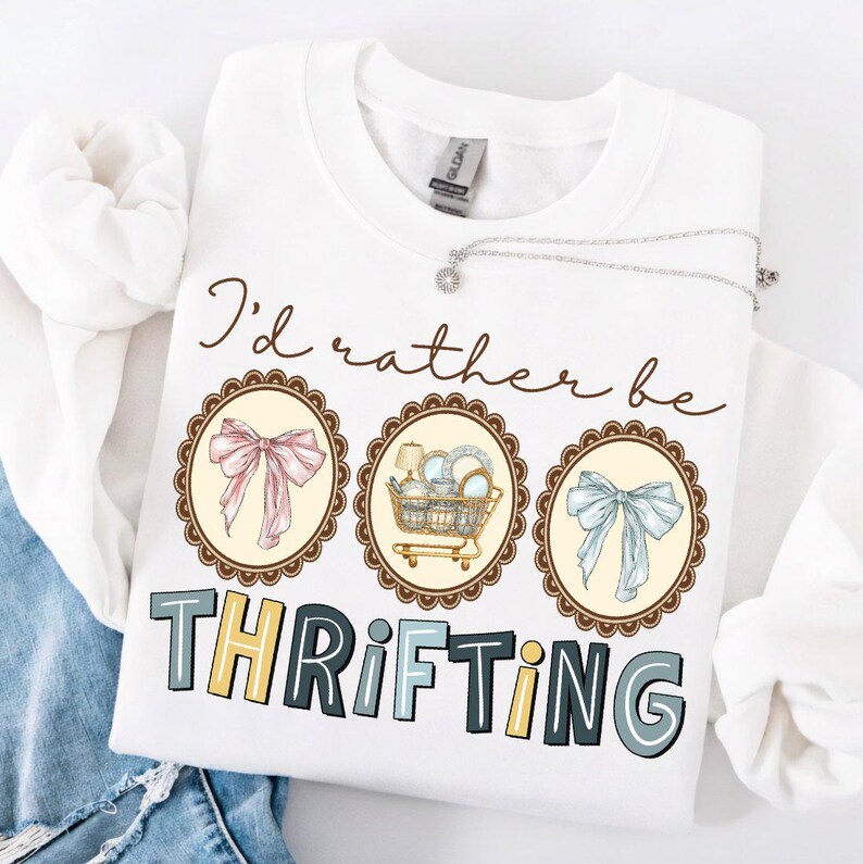 May include: White sweatshirt with the text "I'd rather be thrifting." The design includes three circular illustrations: a pink bow, a shopping cart with dishes, and a blue bow. The word "THRIFTING" is in colorful block letters.