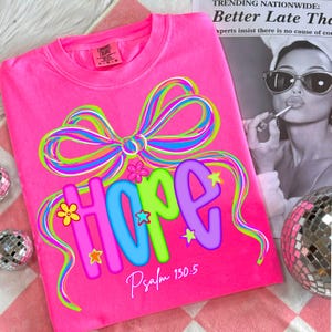 Puede incluir: Una camiseta rosa brillante con la palabra "HOPE" en letras coloridas, adornada con un gran lazo y detalles florales. El texto "Salmo 130:5" est&aacute; impreso debajo. La camiseta es de la marca Comfort Colors.