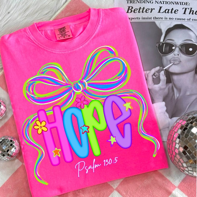 Puede incluir: Una camiseta rosa brillante con la palabra "HOPE" en letras coloridas, adornada con un gran lazo y detalles florales. El texto "Salmo 130:5" est&aacute; impreso debajo. La camiseta es de la marca Comfort Colors.