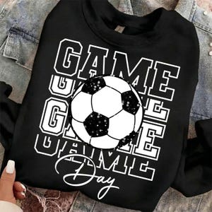 Może przedstawiać: Czarna bluza z napisem "GAME DAY" w białych literach i grafiką piłki nożnej. Piłka nożna jest czarno-biała z efektem brokatu. Bluza jest na kurtce dżinsowej.