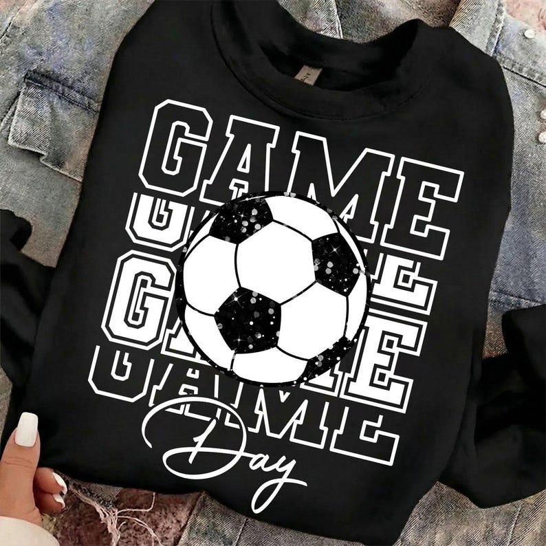 Może przedstawiać: Czarna bluza z napisem "GAME DAY" w białych literach i grafiką piłki nożnej. Piłka nożna jest czarno-biała z efektem brokatu. Bluza jest na kurtce dżinsowej.