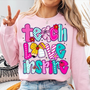 May include: A light pink sweatshirt with the words "Teach Love Inspire" in colorful, patterned letters. The design includes flowers, a pencil, and an apple. The sweatshirt is a comfortable, casual style.