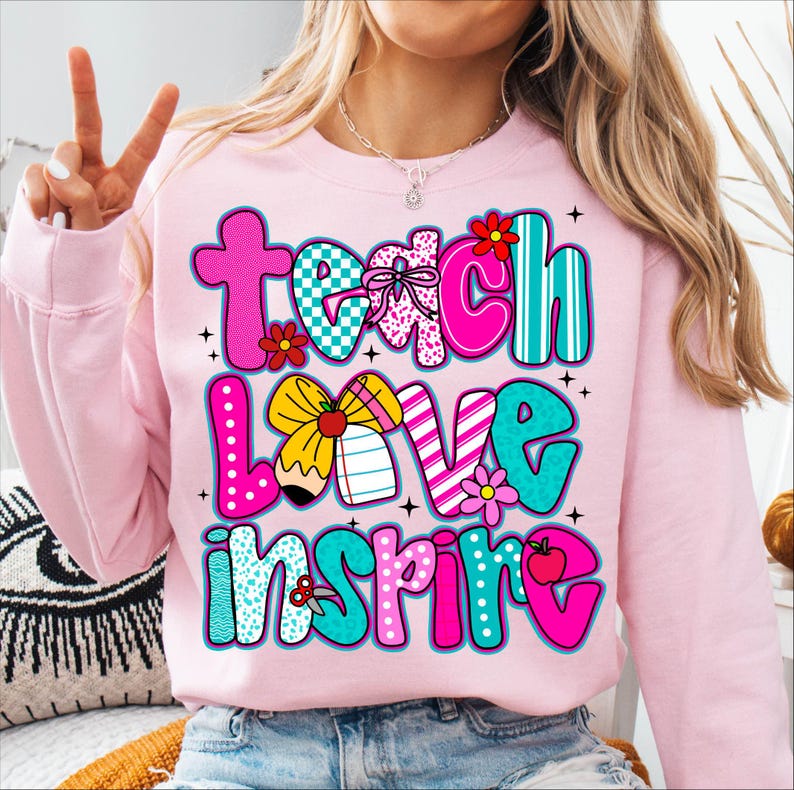 May include: A light pink sweatshirt with the words "Teach Love Inspire" in colorful, patterned letters. The design includes flowers, a pencil, and an apple. The sweatshirt is a comfortable, casual style.