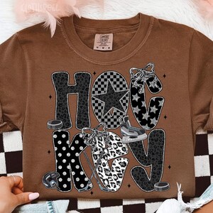 Peut inclure: T-shirt marron avec le mot "HOCKEY" dans un m&eacute;lange de motifs, dont l'imprim&eacute; l&eacute;opard, des &eacute;toiles et un motif &agrave; carreaux. Le design comprend des palets de hockey, un casque et des patins &agrave; glace.