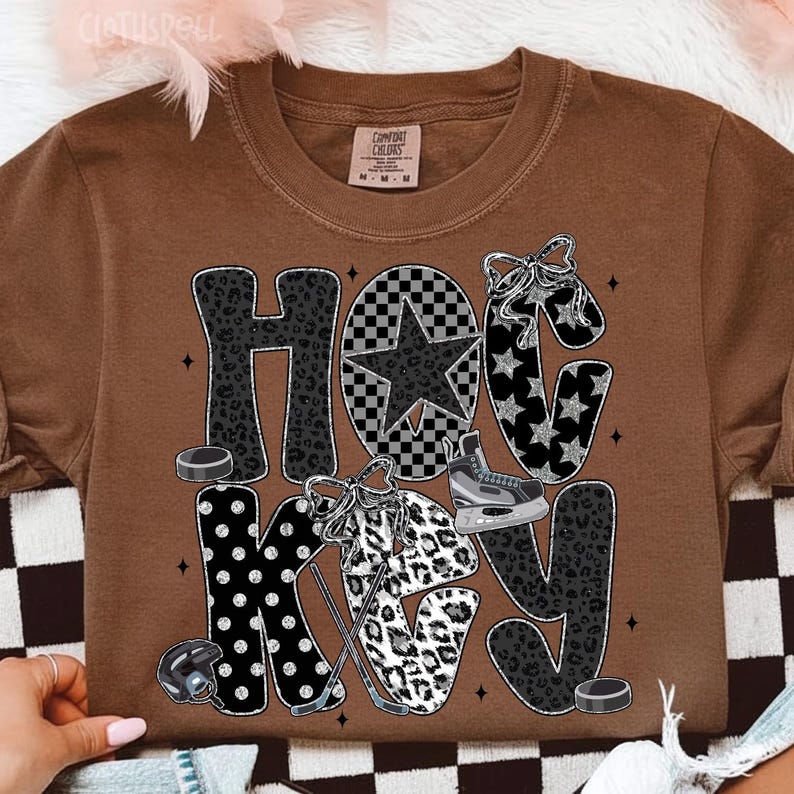 Peut inclure: T-shirt marron avec le mot "HOCKEY" dans un m&eacute;lange de motifs, dont l'imprim&eacute; l&eacute;opard, des &eacute;toiles et un motif &agrave; carreaux. Le design comprend des palets de hockey, un casque et des patins &agrave; glace.