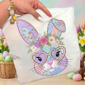 Puede incluir: Un coj&iacute;n decorativo de Pascua con un dise&ntilde;o de conejo de patchwork. El conejo tiene gafas en forma de coraz&oacute;n y detalles florales, en tonos pastel de rosa, morado y azul. El coj&iacute;n est&aacute; sostenido por una mano con una pulsera dorada.