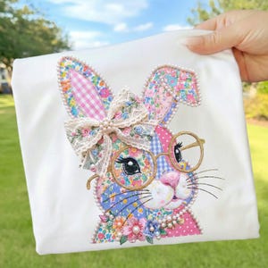 May include: White fabric featuring an embroidered bunny design. The bunny has floral patterned ears, a pink nose, and wears round glasses. A lace bow with pearls adorns the bunny's head, and a pearl border outlines the design. The bunny is surrounded by colorful flowers.