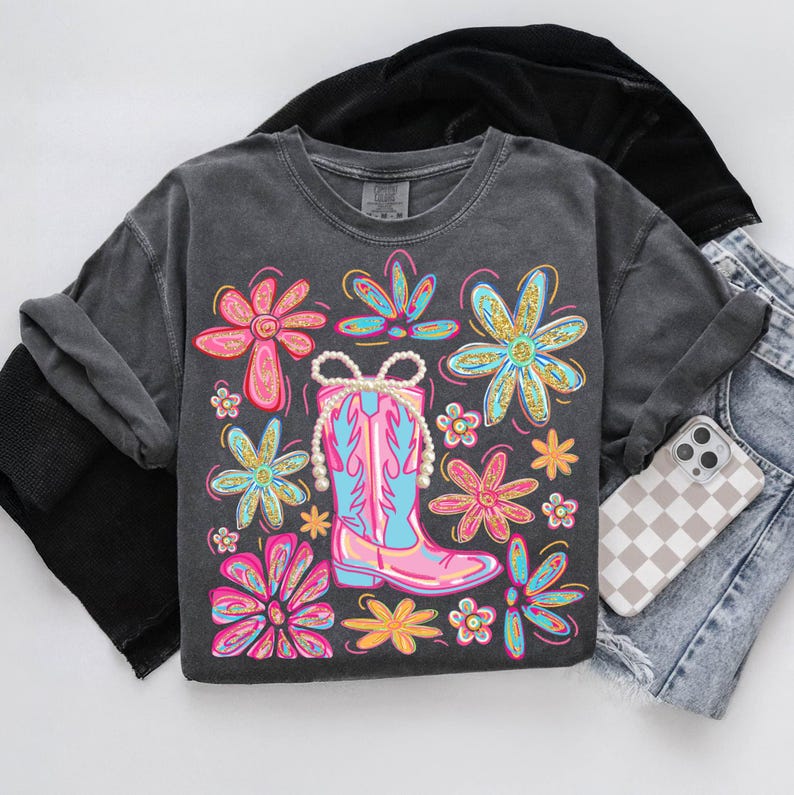 May include: Dark gray t-shirt featuring a colorful graphic of a pink and blue cowboy boot adorned with pearls, surrounded by various flowers in pink, blue, yellow, and orange. The shirt is styled with denim shorts and a phone case.