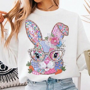 May include: White sweatshirt featuring a colorful bunny design. The bunny has floral patterned ears, a pink nose, and is wearing jeweled glasses. The design includes flowers and carrots, creating a festive and whimsical look.
