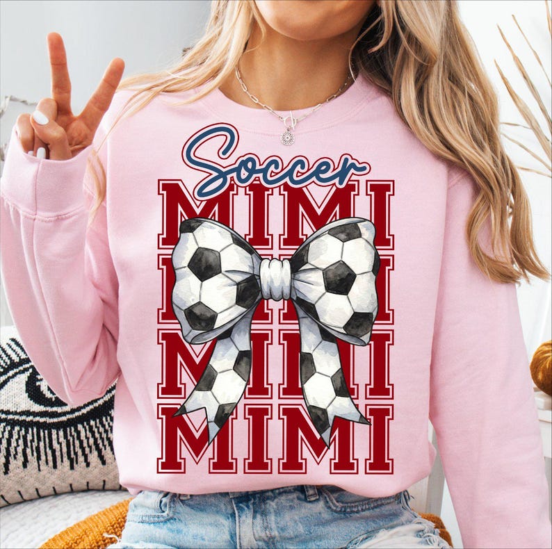 May include: A light pink sweatshirt with the text "Soccer MIMI" in red and blue. The design features a bow made of soccer balls. The sweatshirt is a comfortable, casual style.