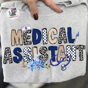 Puede incluir: Una sudadera gris claro con las palabras "MEDICAL ASSISTANT" en una mezcla de azul, beige y estampado de leopardo. El diseño incluye un gorro de enfermera, estetoscopio, jeringa y detalles florales azules. El logotipo de la marca "clothspell" está en la esquina superior izquierda.