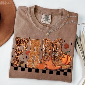 Peut inclure: T-shirt marron avec le mot "FALL" en imprim&eacute; l&eacute;opard et paillettes. Le t-shirt pr&eacute;sente un motif sur le th&egrave;me de l'automne avec des bottes de cow-boy, des citrouilles et un gland. Un collier argent&eacute; est drap&eacute; sur le t-shirt.