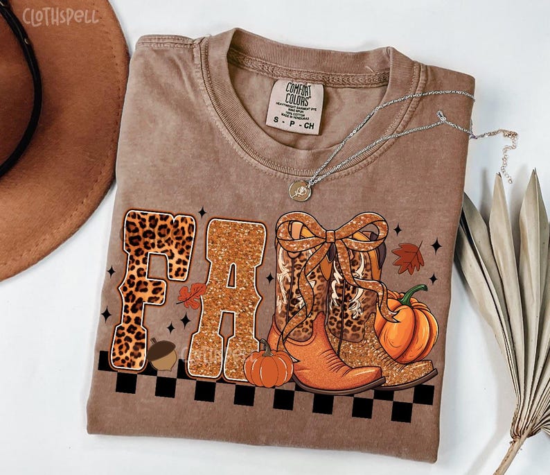 Peut inclure: T-shirt marron avec le mot "FALL" en imprim&eacute; l&eacute;opard et paillettes. Le t-shirt pr&eacute;sente un motif sur le th&egrave;me de l'automne avec des bottes de cow-boy, des citrouilles et un gland. Un collier argent&eacute; est drap&eacute; sur le t-shirt.