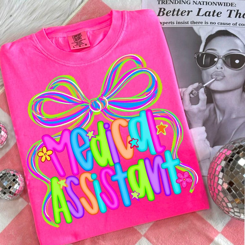 Puede incluir: Una sudadera rosa brillante con las palabras "Medical Assistant" en letras coloridas. Un gran lazo multicolor adorna la parte superior del texto. La camiseta est&aacute; sobre una superficie a cuadros rosa y blanco.