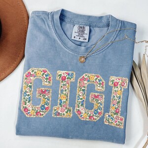 K&ouml;nnte beinhalten: Ein hellblaues Comfort Colors T-Shirt mit dem Namen "GIGI" in Buchstaben mit floralem Druck. Eine goldene Halskette mit einem kleinen Anh&auml;nger liegt auf dem Shirt. Ein brauner Hut und getrocknete Palmbl&auml;tter sind im Hintergrund.