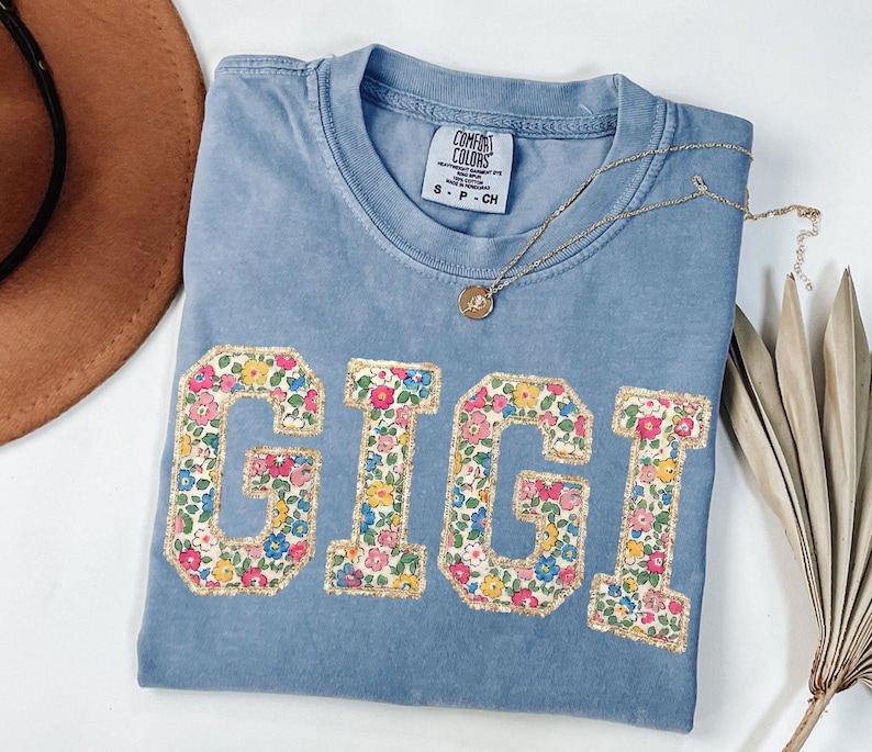 K&ouml;nnte beinhalten: Ein hellblaues Comfort Colors T-Shirt mit dem Namen "GIGI" in Buchstaben mit floralem Druck. Eine goldene Halskette mit einem kleinen Anh&auml;nger liegt auf dem Shirt. Ein brauner Hut und getrocknete Palmbl&auml;tter sind im Hintergrund.