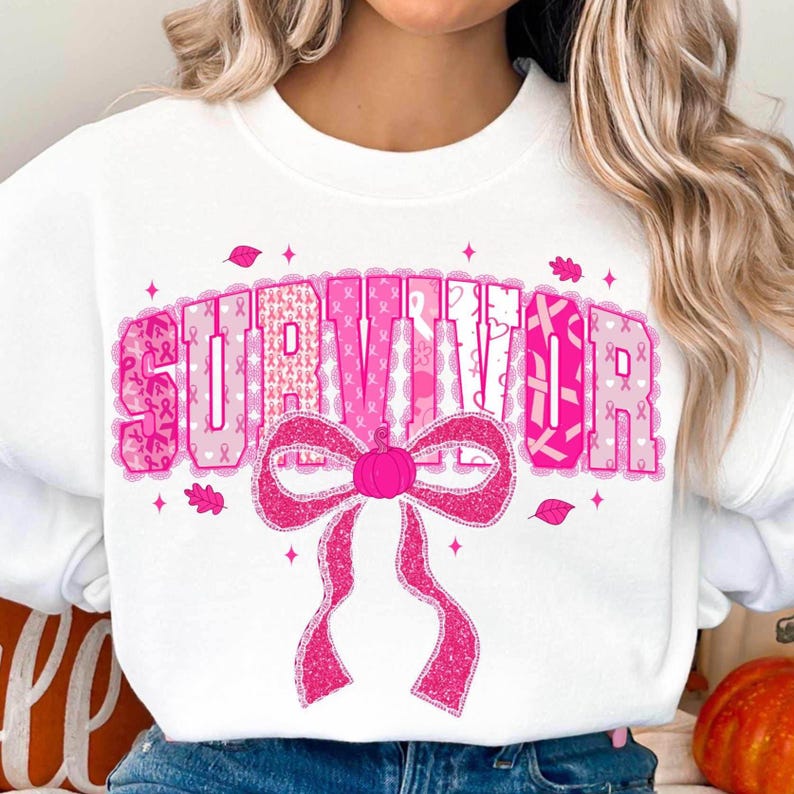 May include: White sweatshirt with the word "SURVIVOR" in pink, decorated with pink ribbons and a bow with a pumpkin center. The design includes pink leaves and small stars, creating a fall-themed aesthetic.