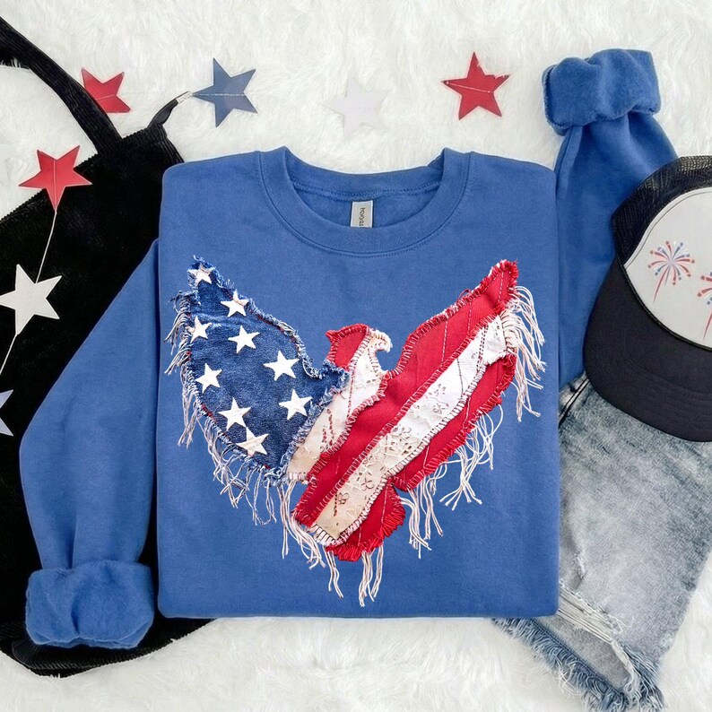 May include: A blue crewneck sweatshirt with a patriotic eagle design. The eagle's wings are made of denim with white stars and red and white striped fabric. The sweatshirt is paired with denim shorts and a black trucker hat.