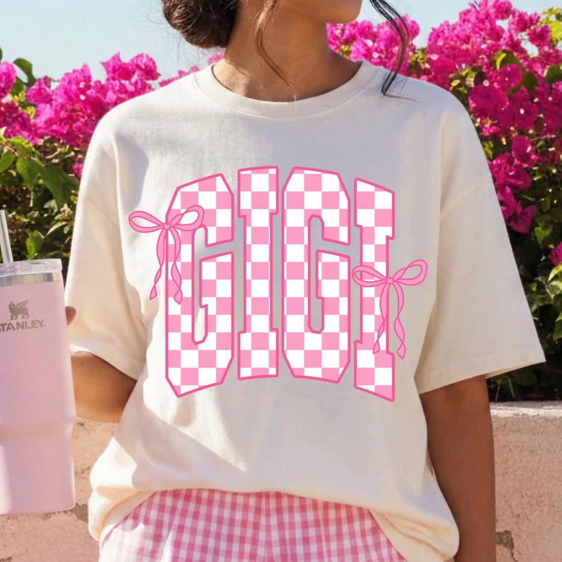 May include: Cream-colored t-shirt with the word "GIGI" in a pink and white checkered pattern. The letters are outlined in pink and adorned with pink bows. A pink Stanley tumbler is visible on the left.