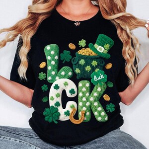 May include: Black t-shirt with a St. Patrick's Day design. The word "LUCK" is spelled out in green and white textured letters, with shamrocks, a pot of gold, and a green hat. Gold coins and a horseshoe are also featured.