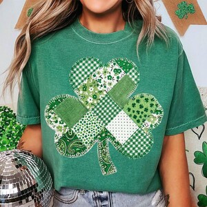 May include: Green t-shirt featuring a patchwork shamrock design. The shamrock is composed of various green fabrics with different patterns, including gingham, floral, and paisley. The shirt is a solid green color.