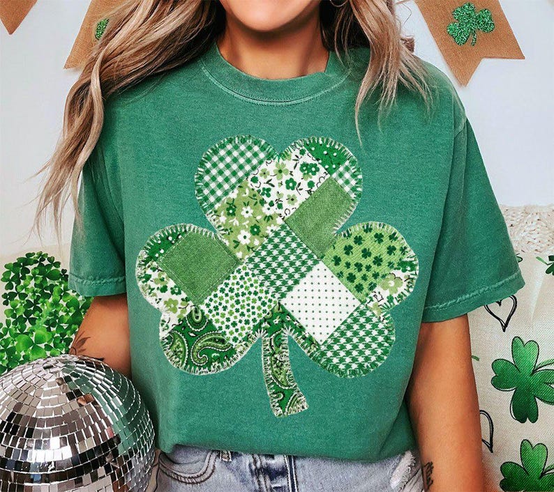 May include: Green t-shirt featuring a patchwork shamrock design. The shamrock is composed of various green fabrics with different patterns, including gingham, floral, and paisley. The shirt is a solid green color.