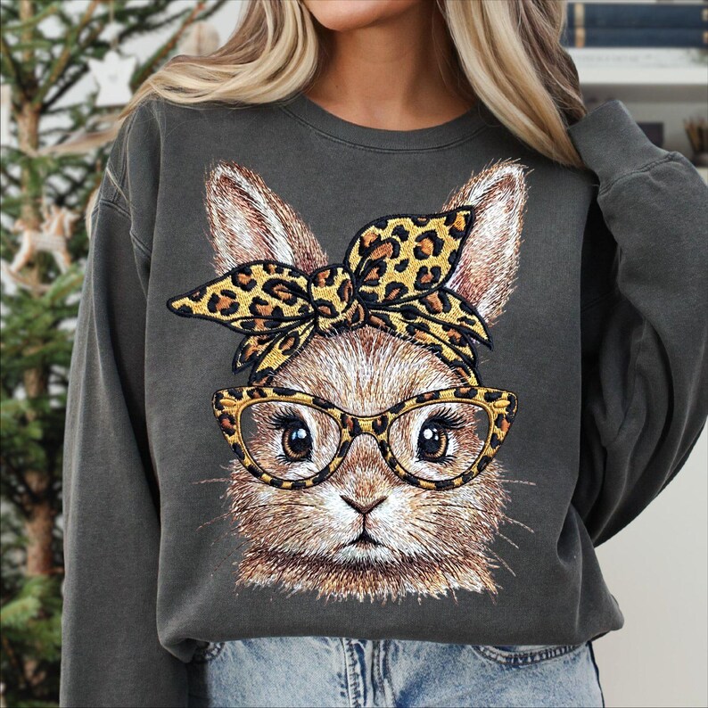 May include: Dark gray sweatshirt featuring a detailed illustration of a bunny wearing glasses and a leopard print bow. The bunny's face is centered, with large eyes and a detailed fur pattern. The sweatshirt has long sleeves and a crew neck.