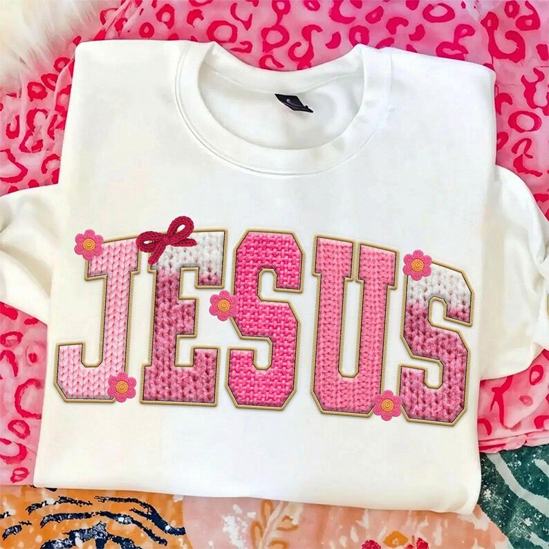 May include: White sweatshirt with the word "JESUS" in pink, knitted-style letters outlined in gold. A small red bow and pink flowers decorate the top of the letters. The sweatshirt is on a pink and white patterned surface.