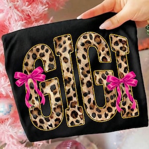 May include: Black t-shirt with the word "GIGI" in a leopard print design outlined in gold. Two pink bows are attached to the shirt. The shirt is held by a person.