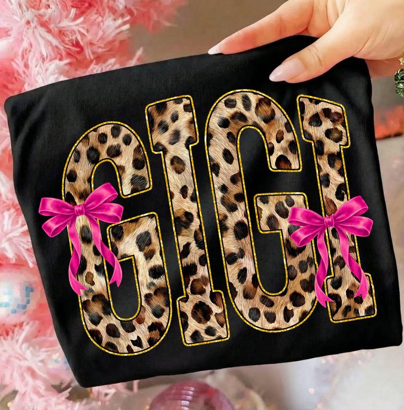 May include: Black t-shirt with the word "GIGI" in a leopard print design outlined in gold. Two pink bows are attached to the shirt. The shirt is held by a person.