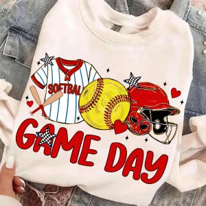 May include: White sweatshirt with a softball design. It features a yellow softball, red helmet, and a baseball jersey graphic. The words "GAME DAY" are in red, with star and heart accents. A fun design for sports fans.
