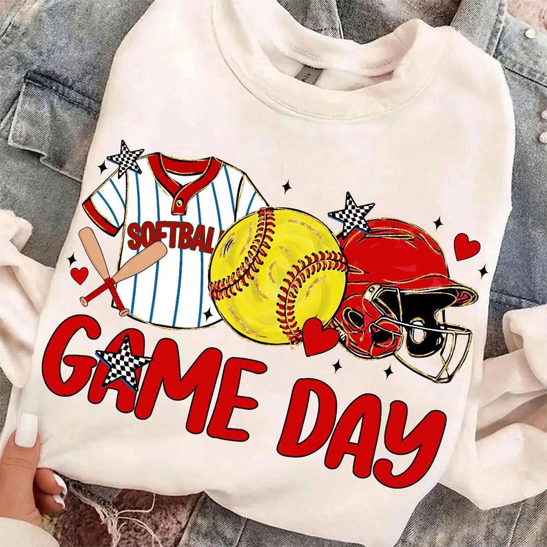 May include: White sweatshirt with a softball design. It features a yellow softball, red helmet, and a baseball jersey graphic. The words "GAME DAY" are in red, with star and heart accents. A fun design for sports fans.