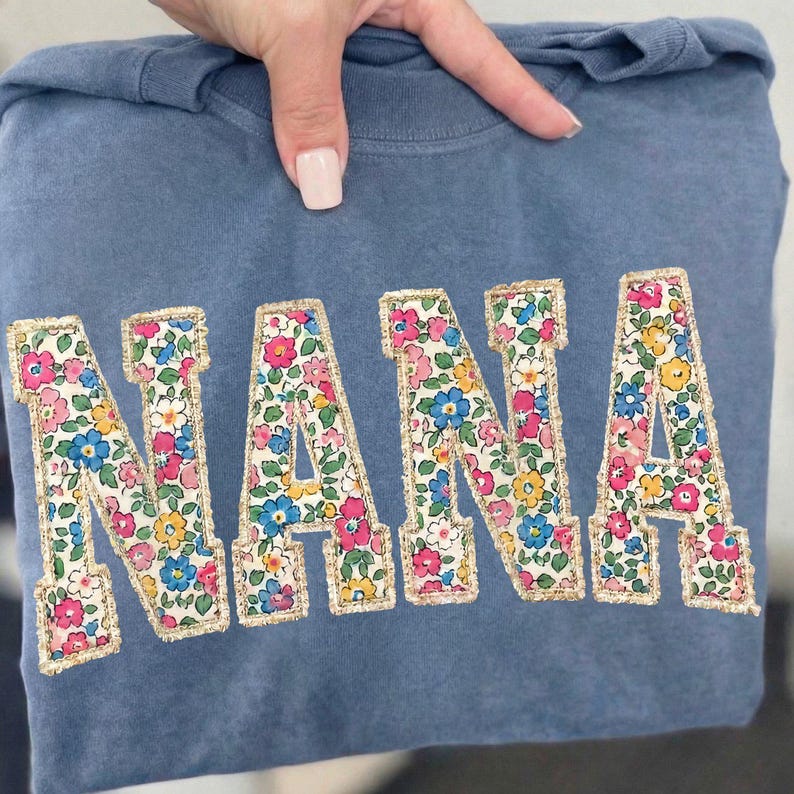 K&ouml;nnte beinhalten: Blaues Sweatshirt mit dem Wort "NANA" auf der Vorderseite gestickt. Die Buchstaben sind mit einem Blumenmuster in Rosa, Blau, Gelb und Wei&szlig; gef&uuml;llt und mit goldenen Zierleisten versehen.