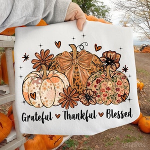 May include: White sweatshirt featuring a fall-themed design with pumpkins, flowers, and hearts. The pumpkins have floral and leopard print patterns. The text "Grateful Thankful Blessed" is printed at the bottom.