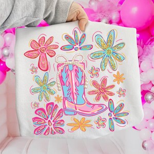 May include: White sweatshirt featuring a colorful illustration of a pink and blue cowboy boot with a pearl necklace, surrounded by pink, blue, and yellow flowers with gold glitter accents. The design is held by a person's hand.