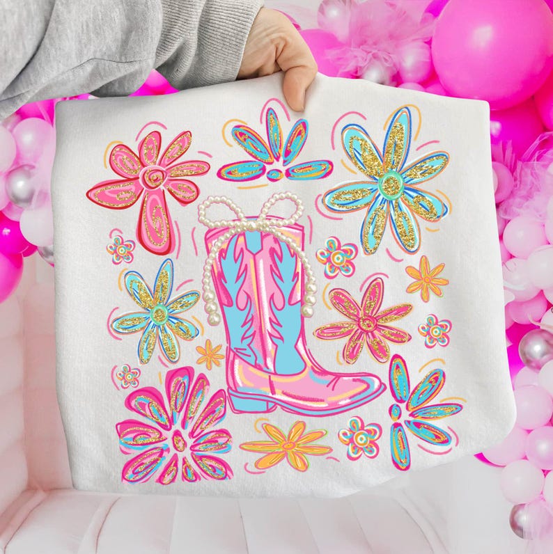 May include: White sweatshirt featuring a colorful illustration of a pink and blue cowboy boot with a pearl necklace, surrounded by pink, blue, and yellow flowers with gold glitter accents. The design is held by a person's hand.