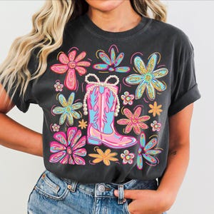 May include: Dark gray t-shirt featuring a colorful graphic of a pink and blue cowboy boot adorned with a pearl necklace, surrounded by vibrant flowers in pink, blue, and yellow. The design has a distressed, vintage look.