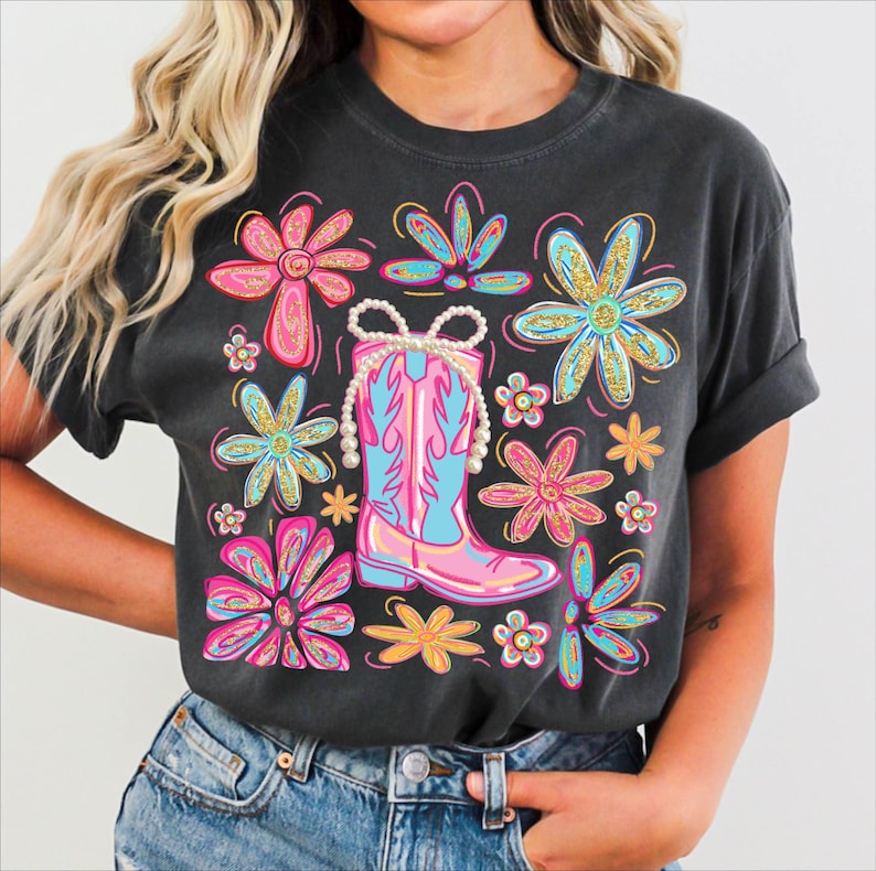 May include: Dark gray t-shirt featuring a colorful graphic of a pink and blue cowboy boot adorned with a pearl necklace, surrounded by vibrant flowers in pink, blue, and yellow. The design has a distressed, vintage look.