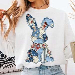 May include: White sweatshirt featuring a patchwork bunny design. The bunny is made of blue fabric with floral, polka dot, and striped patterns in red, white, and blue. The sweatshirt is a casual, comfortable style.